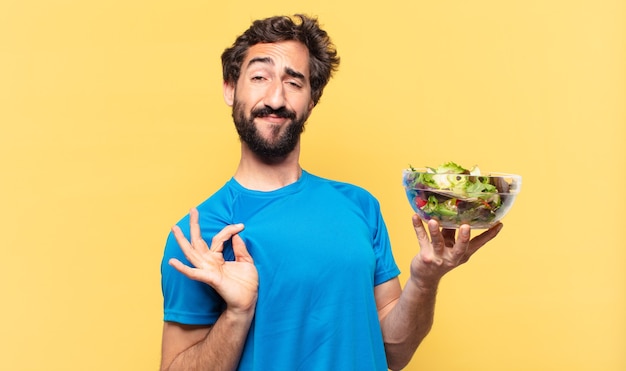 Joven atleta barbudo loco con expresión feliz con ensalada