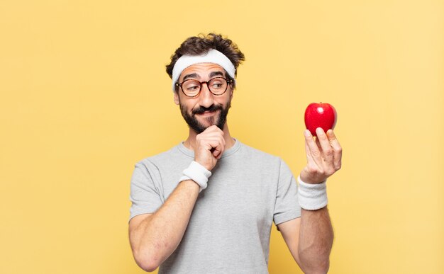 Joven atleta barbudo loco pensando expresión y sosteniendo una manzana