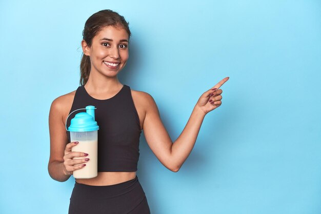 Joven atleta con batido de proteínas listo para el gimnasio sonriendo y señalando a un lado mostrando algo