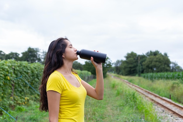 Joven atleta femenina étnica bebiendo agua después de entrenar en la naturaleza