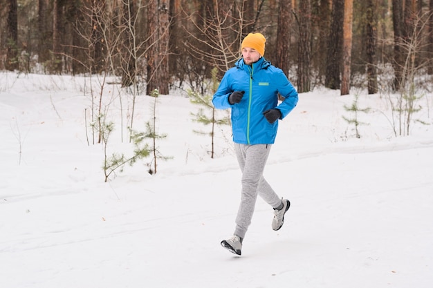 Joven atleta en gorro corriendo por el camino nevado en el bosque de invierno mientras entrena solo