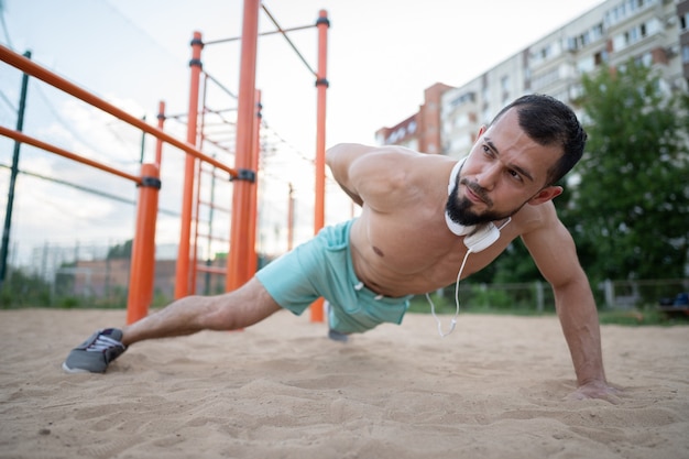 Un joven atleta hace flexiones con una mano en la arena en verano. Deportes, fitness, concepto de entrenamiento en la calle.