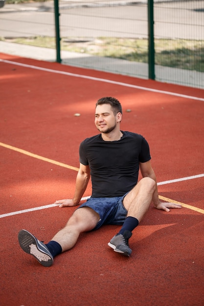 Un joven atleta masculino con barba se sienta en una cancha de tenis roja. Chico guapo en camiseta y pantalones cortos