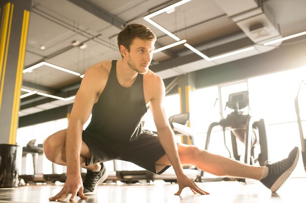 Joven atleta masculino en camiseta deportiva y pantalones cortos flexionando las piernas durante el entrenamiento deportivo en el gimnasio