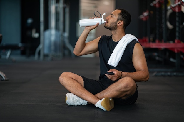 Joven atleta masculino negro bebiendo agua mientras se relaja después de entrenar en el gimnasio