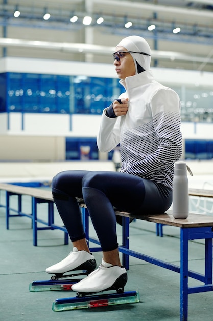 Joven atleta de patinaje de velocidad de pista corta en uniforme y patinadores sentados en un banco