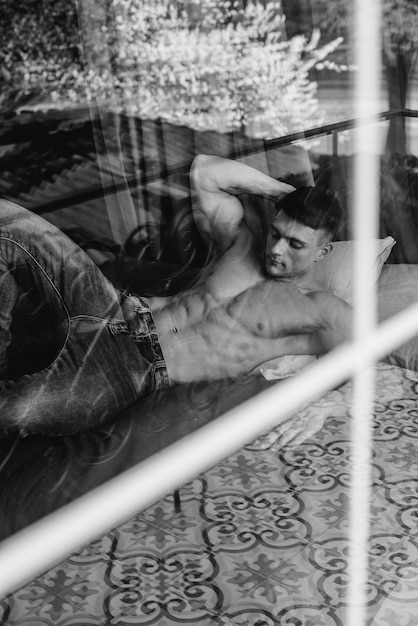 Un joven atleta sexy con abdominales perfectos está acostado en la cama en el estudio en topless en jeans. Estilo de vida saludable, nutrición adecuada, programas de entrenamiento y nutrición para adelgazar. En blanco y negro.