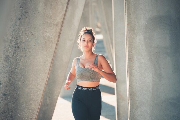 Joven atlética afroamericana, peinado rizado, corriendo al aire libre en la ciudad, corredor Deportes urbanos en la ciudad. Hora del atardecer con sombras. Mujer seria concentrándose en el tren de competición.