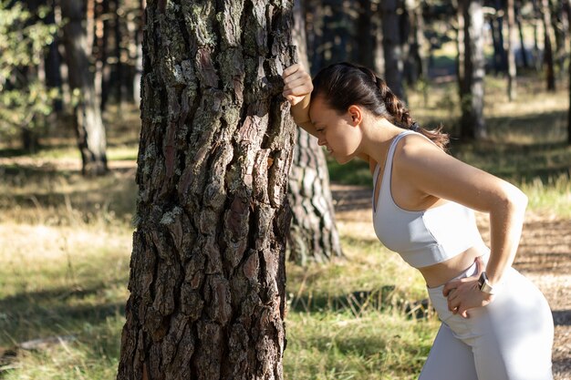 Joven atlética con aspecto cansado después de trotar o hacer ejercicio, apoyado contra un árbol