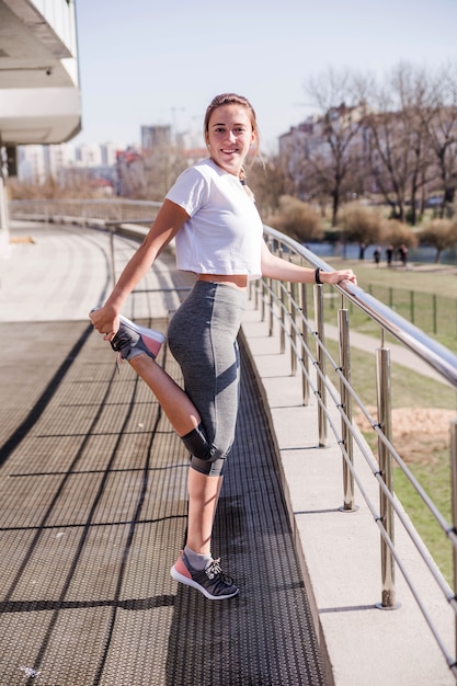 Joven atlética en una camiseta blanca y leggings grises antes de un entrenamiento al aire libre hace un entrenamiento. El clima es agradable, soleado