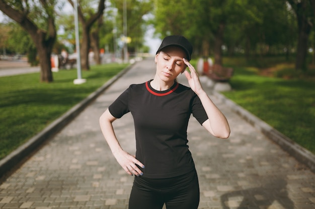 Joven atlética cansada hermosa mujer morena en uniforme negro y gorra de pie, descansando y manteniendo la mano cerca de la cabeza después de correr, entrenando en el parque de la ciudad al aire libre