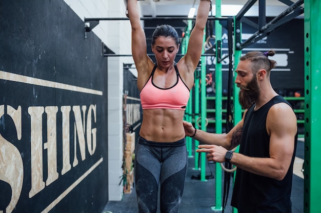 Joven atlética haciendo pull up en el bar de gimnasia ayudado por entrenador personal