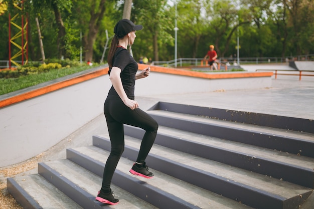 Joven atlética hermosa mujer morena en uniforme negro, gorra con auriculares haciendo ejercicios deportivos, entrenando y corriendo, subiendo escaleras en el parque de la ciudad al aire libre