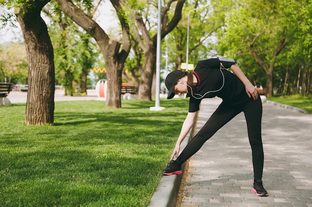 Foto joven atlética hermosa mujer morena en uniforme negro y gorra con auriculares haciendo ejercicios de estiramiento deportivo, calentamiento antes de correr en el parque de la ciudad al aire libre