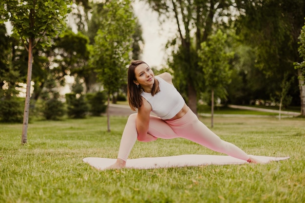 Joven atlética practica yoga en colchoneta gimnástica en un parque verde sobre césped Haz ejercicios de estiramiento de piernas y espalda en el fondo de la naturaleza de ropa deportiva Concepto de estilo de vida saludable y armonía con el cuerpo