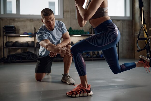 Joven atlética con ropa deportiva entrenando piernas con correas de suspensión bajo el control del entrenador. Entrenador profesional enseñando al cliente a ponerse en cuclillas