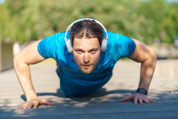 Joven atlético en auriculares haciendo flexiones mientras hace ejercicio al aire libre