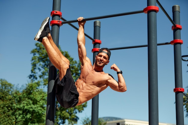 Joven atlético colgando de las barras en el gimnasio de calistenia al aire  libre sonriendo