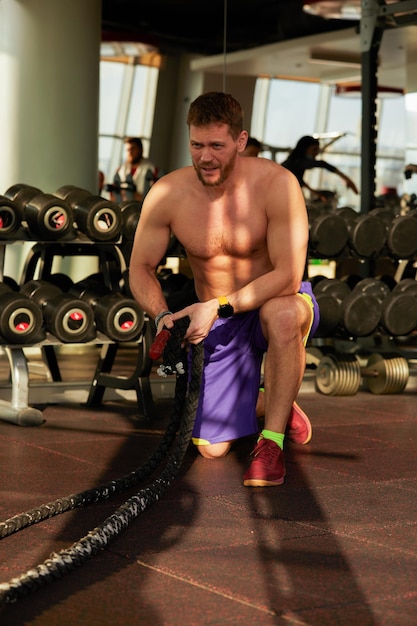 Joven atlético con cuerda de batalla haciendo ejercicio en entrenamiento funcional gimnasio