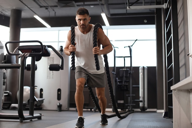 Joven atlético con cuerda de batalla haciendo ejercicio en el gimnasio de entrenamiento funcional.