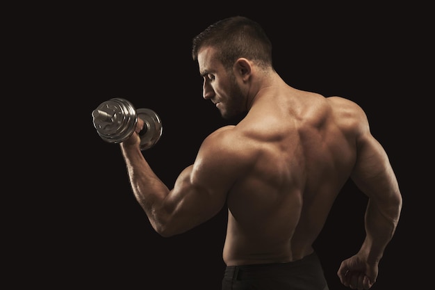 Joven atlético con mancuernas sobre fondo negro, haciendo fuertes ejercicios de bíceps. Torso desnudo, cuerpo musculoso, concepto de culturismo, espacio de copia