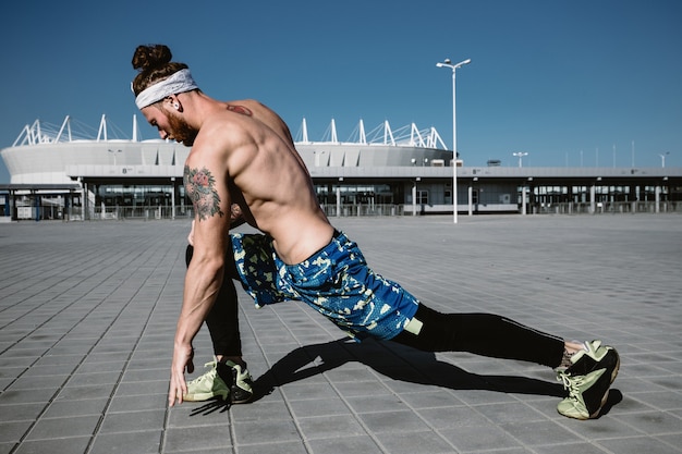 Joven atlético con un torso desnudo con diadema vestido con leggings negros y pantalones cortos azules está haciendo estiramientos en losas de pavimentación en el fondo del estadio en un día cálido y soleado.