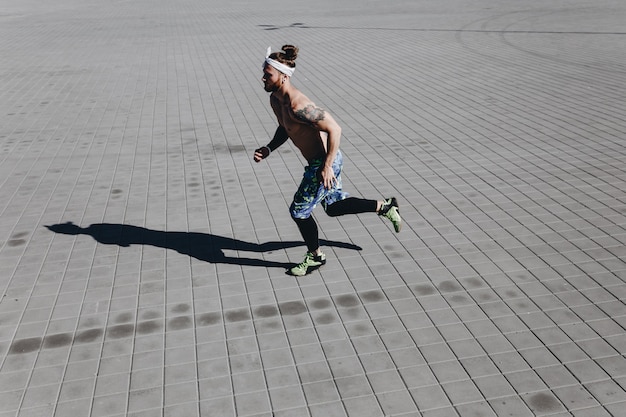 Joven atlético con un torso desnudo con tatuajes y diadema en la cabeza vestido con leggings negros y pantalones cortos azules se ejecuta en losas de pavimentación en un día cálido y soleado.