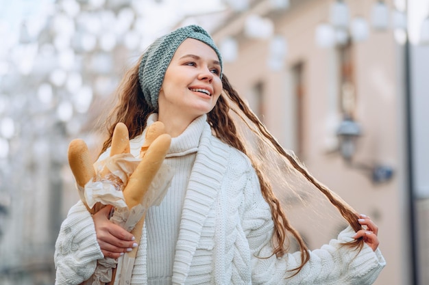 Una joven y atractiva belleza feliz con cabello largo sonríe brillantemente y toca un mechón de cabello. Sosteniendo unos panes en la mano. Campaña publicitaria de productos de panadería. Espacio para texto o logo.