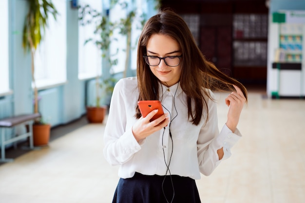 Joven atractiva chica caucásica con teléfono móvil y auriculares de pie dentro de la universidad o escuela