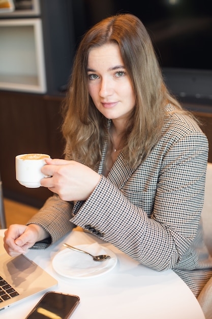 Una joven y atractiva chica regordeta está sentada en un café tomando café y trabajando en una computadora portátil con fotos de alta calidad