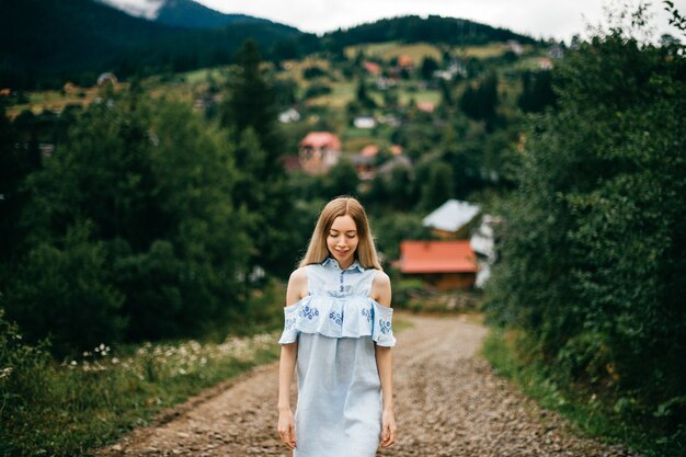 Joven atractiva chica rubia elegante en vestido azul posando en la carretera en el campo