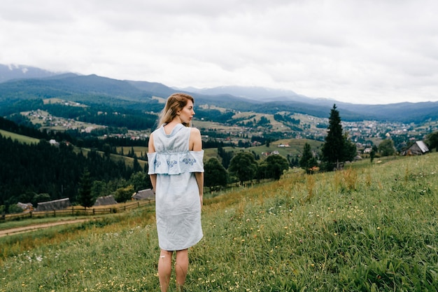 Joven atractiva chica rubia elegante en vestido azul posando sobre un paisaje pintoresco