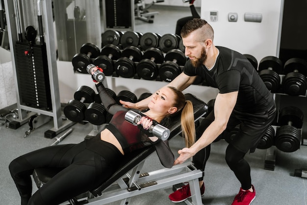 Joven atractiva chica rubia en forma haciendo ejercicio con mancuernas con la ayuda de su entrenador