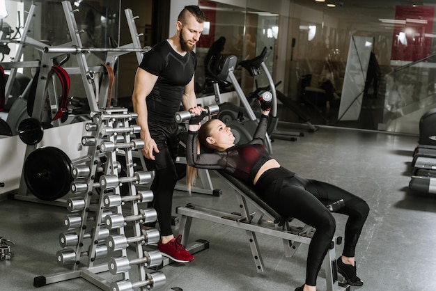 Joven atractiva chica rubia en forma haciendo ejercicio con mancuernas con la ayuda de su entrenador