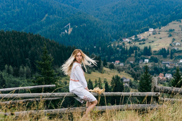 Foto joven atractiva chica rubia en vestido blanco con adorno sentado en la valla de madera con ramo de espiguillas sobre el pintoresco paisaje de la campiña