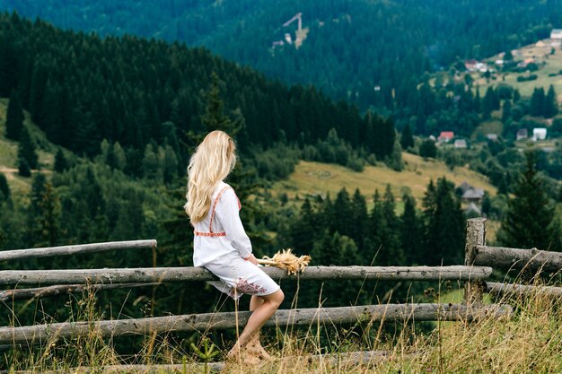 Joven atractiva chica rubia en vestido blanco con adorno sentado en la valla de madera con ramo de espiguillas sobre el pintoresco paisaje rural
