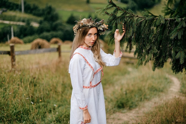 Joven atractiva chica rubia en vestido blanco con bordado y corona de flores en la cabeza posando con rama de abeto sobre paisaje de campo