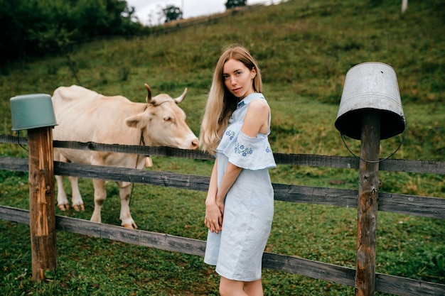 Joven atractiva elegante rubia en vestido azul romántico posando con vaca en el campo