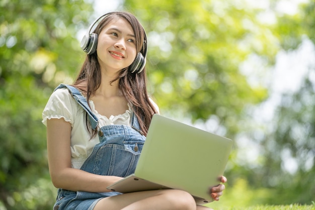 Joven y atractiva empleada asiática usa auriculares para escuchar el estudio de desarrollo de cursos digitales en línea en el parque