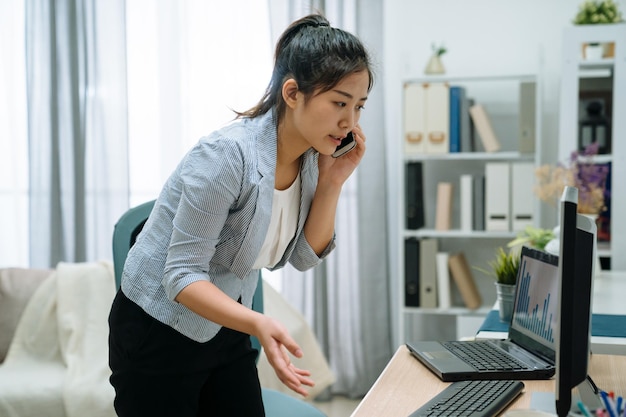 Joven y atractiva empresaria asiática japonesa hablando por teléfono móvil mientras está de pie en el escritorio en una oficina en casa brillante y de alta clave. mujer elegante gerente discutiendo sobre el teléfono móvil en el día tim