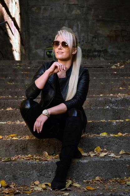 Joven atractiva y hermosa mujer rubia con gafas de sol sentada en las escaleras de piedra y sonriendo