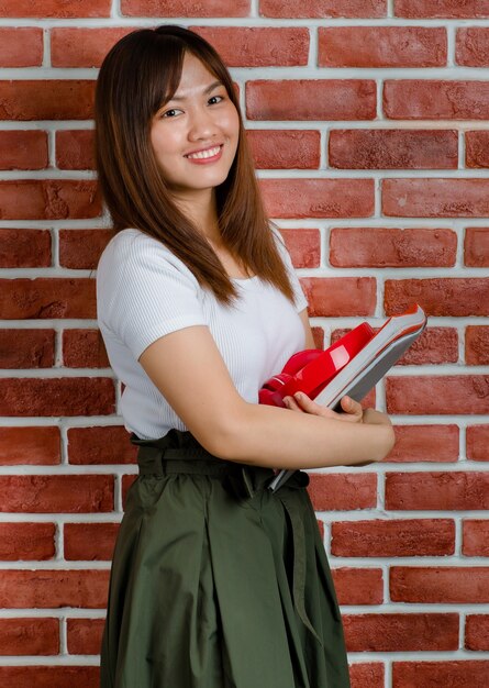 Joven atractiva mujer asiática en camiseta blanca y falda verde de pie y sosteniendo carpetas y cuaderno sobre fondo de pared de ladrillo naranja.