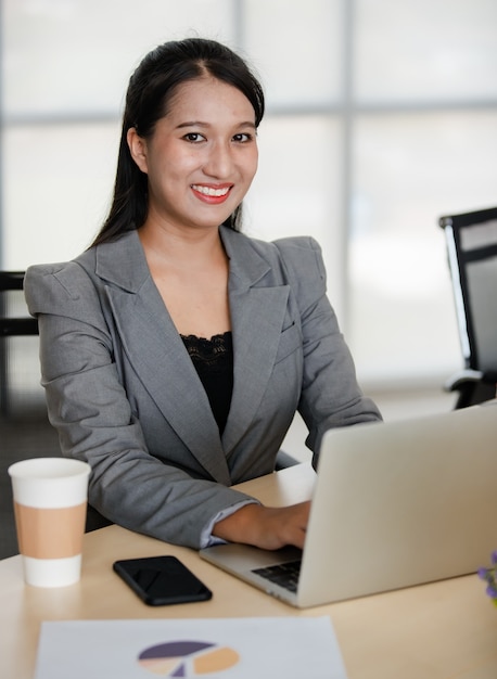 Joven atractiva mujer asiática en traje gris trabajando en equipo portátil en la oficina de aspecto moderno. Concepto de estilo de vida de oficina moderna.