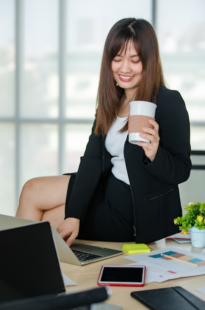 Joven atractiva mujer asiática en traje negro trabajando en la computadora portátil mientras toma café en la oficina de aspecto moderno con fondo de ventanas borrosas. Concepto de estilo de vida de oficina moderna.