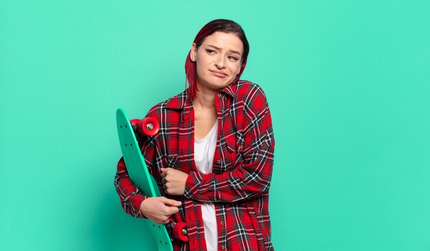 Joven atractiva mujer de cabello rojo encogiéndose de hombros, sintiéndose confundida e insegura, dudando con los brazos cruzados y la mirada perpleja y sosteniendo una patineta