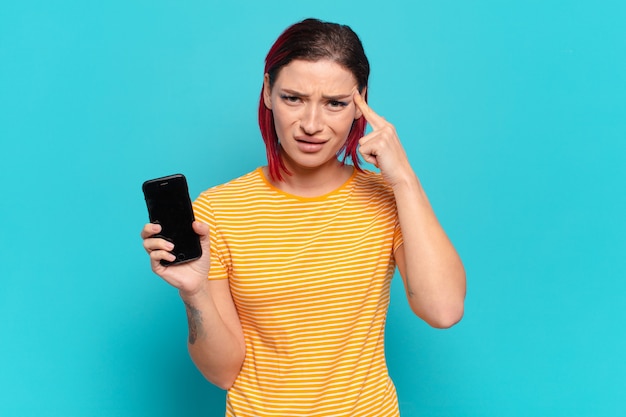 Joven atractiva mujer de cabello rojo que se siente confundida y desconcertada, mostrando que estás loco, loco o fuera de tu mente y mostrando su celular