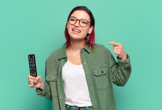 Joven atractiva mujer de cabello rojo sonriendo con confianza apuntando a su propia sonrisa amplia, actitud positiva, relajada y satisfecha y sosteniendo un control remoto de tv