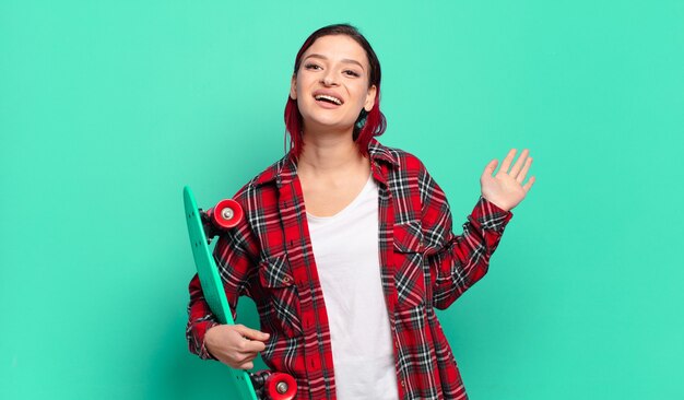 Foto joven atractiva mujer de cabello rojo sonriendo feliz y alegremente, saludando con la mano, dándote la bienvenida y saludándote, o diciéndote adiós y sosteniendo una patineta