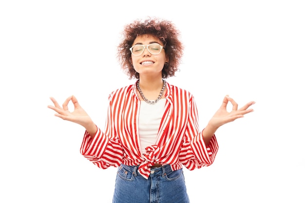 Joven atractiva mujer caucásica rizada con gafas sonriendo en postura meditativa aislada en fondo blanco de estudio haciendo gesto de oración con los dedos mantener la calma y relajarse concepto