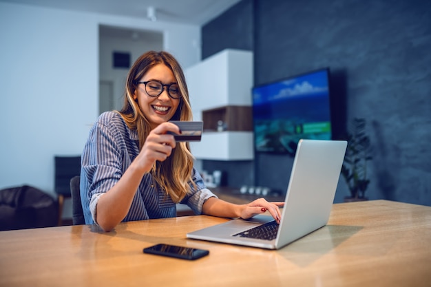 Joven atractiva mujer caucásica sonriente con anteojos con tarjeta de crédito y escribiendo el número de su cuenta bancaria en la computadora portátil mientras está sentado en la mesa de comedor. Concepto de compra en línea.